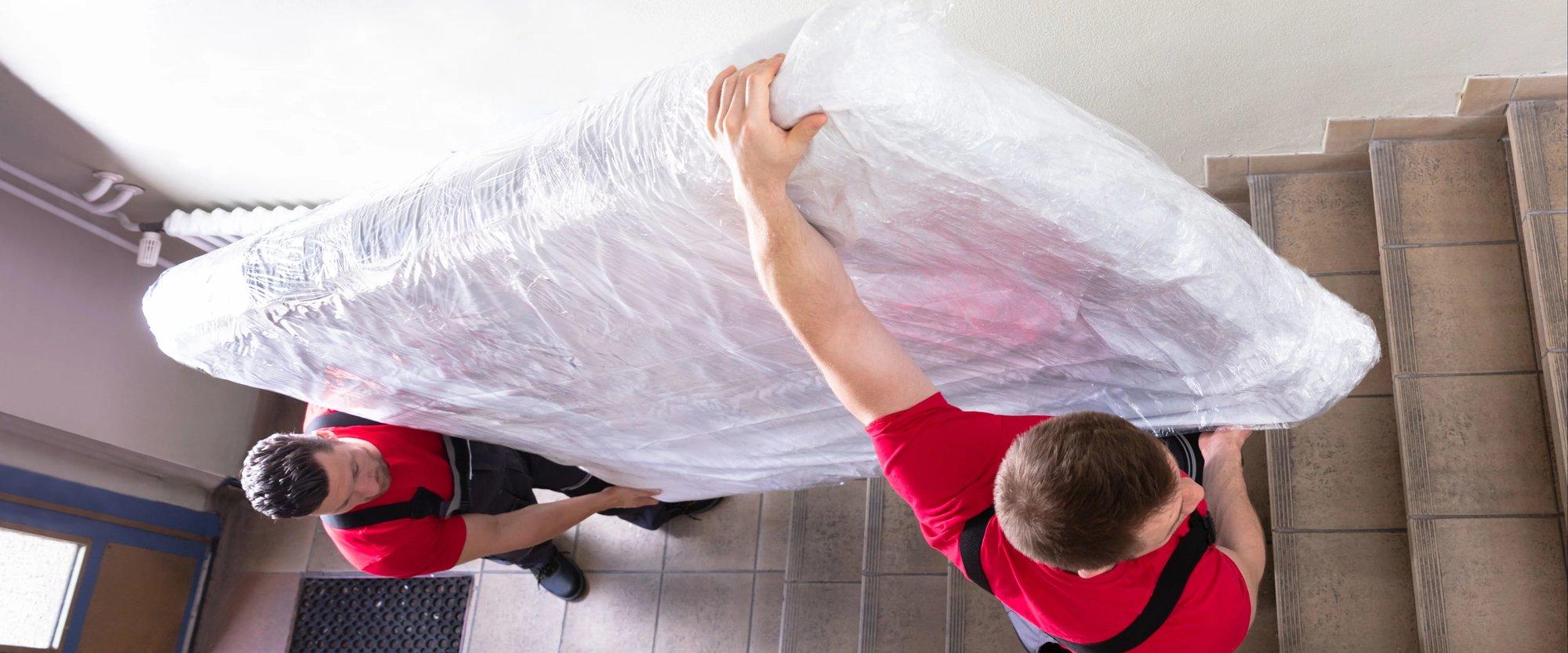 Two men carrying a mattress in a protective cover up a flight of stairs.